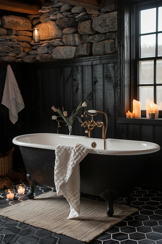 White clawfoot tub with brass faucet against black wood walls and stone, soft white linen towel, woven rug, and candlelight in a cozy dark farmhouse bathroom.
